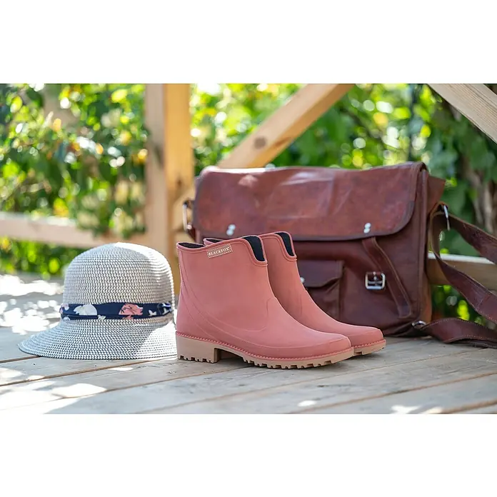 Black Fox Stiefelette Preston - Schuhgröße: 41, Ziegelrot - Ein Paar rosa Blackfox Gummistiefel neben einem Hut und einer braunen Tasche auf einem Holztisch.