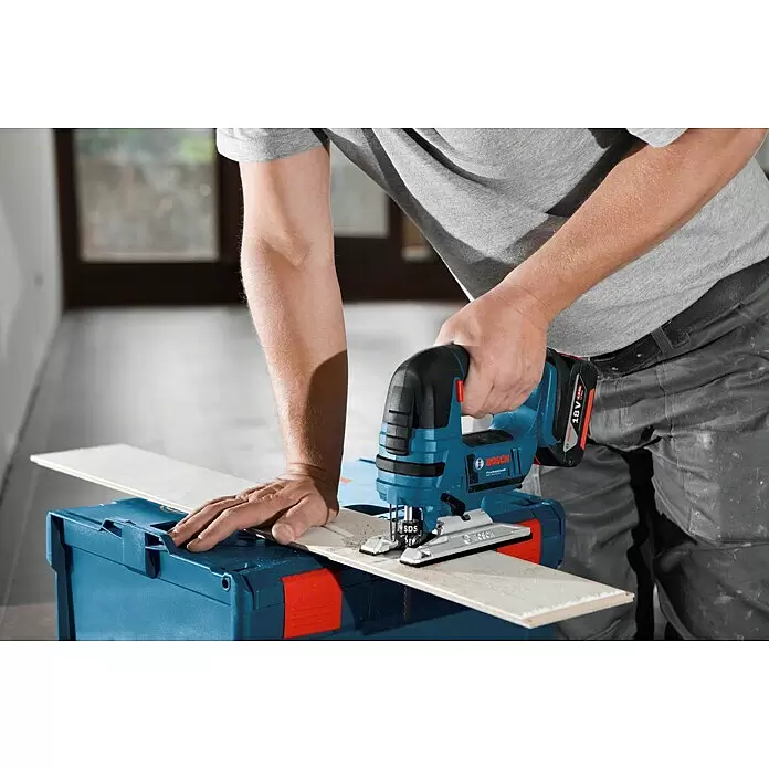 Bosch Professional AMPShare 18V Sierra de calar de batería GST 18V-LI L-Boxx - 18 V, Sin batería, Número de carreras marcha en vacío: 0 /min - 2.700 /min - Persona cortando una tabla de madera clara con una sierra de calar Bosch azul y negra a batería sobre un estuche de herramientas azul.