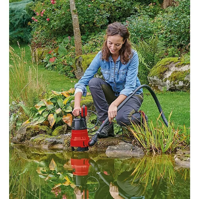 Einhell Bomba para aguas sucias GC-DP 7035 LL - 700 W, Capacidad de bombeo máx.: 16.000 l/h - Una mujer está agachada a la orilla del agua, accionando una bomba sumergible Einhell roja que está sentada y reflejándose en el agua.
