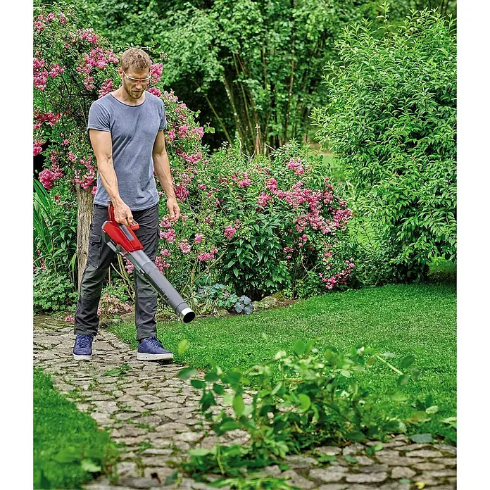Einhell Soplador de hojas de batería TP-LB 18/200 LI - 18 V, 1 batería, Velocidad de soplado: 200 km/h, Potencia de soplado: 670 m³/min - Hombre usando un soplador de hojas en el jardín. El soplador de hojas es rojo y gris. El hombre lleva gafas de seguridad.