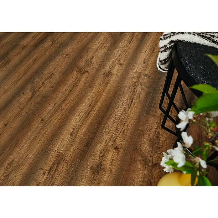 LOGOCLIC Edition Laminat Family Ascoli1.285 x 192 x 7 mm, Landhausdiele, Ascoli - Nahaufnahme eines Bodens mit warmem, braunem Laminat in Holzoptik, ergänzt durch eine elegante schwarze Sitzgelegenheit mit Textildecke.