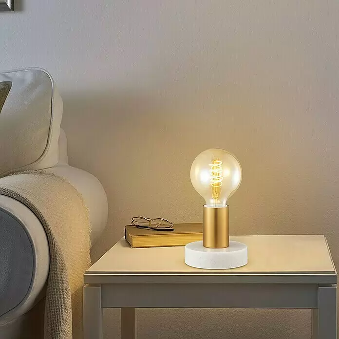 Deko-Tischleuchte Sten - 10 W, L x B x H: 10 x 9 x 9 cm, Gold - Eine kleine Tischlampe mit weißem Marmorsockel, goldenem Stiel und leuchtender Glühfadenbirne auf einem Nachttisch.