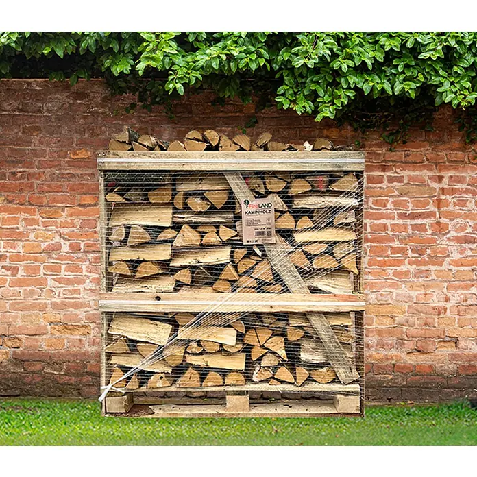 Fireland Kaminholz Raummeter - Buche, Länge Holzscheit: 25 cm - 33 cm - Brennholzpalette vor einer Ziegelwand und üppigem Grün. Das Holz ist sauber gestapelt und mit Netz umwickelt, bereit für den Kamin.
