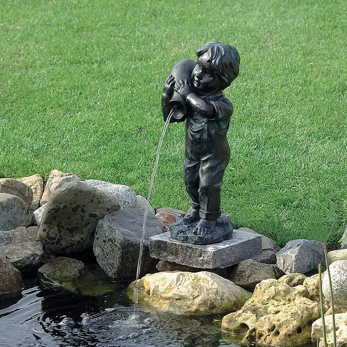 Ubbink Wasserspeier Yannick - Junge - Brunnen-Statue eines Jungen, der einen Krug hält, aus dem Wasser in einen Teich fließt, umgeben von Steinen und üppigem Grün.
