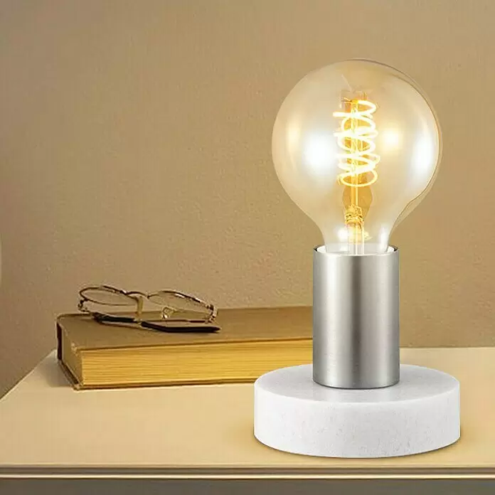 Deko-Tischleuchte Sten - 10 W, L x B x H: 10 x 9 x 9 cm, Stahl matt - Tischlampe mit weißem Marmorsockel, gebürstetem Silberstiel und leuchtender klarer Glühbirne, auf einem Holztisch.
