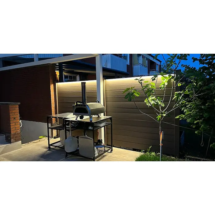 LyghtUp Zaunbeleuchtung Starter-Set - Länge: 173,3 cm, Silber, Passend für: WPC- und Aluminium-Steckzäune, 5 -tlg. - Ein Ooni Pizzaofen steht auf einem schwarzen Metalltisch im Freien, beleuchtet von einer sanften Lichtleiste, die eine gemütliche Atmosphäre schafft.