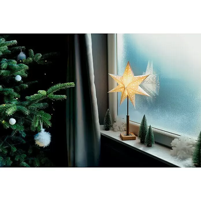 Markslöjd Standstern Solvalla - Ø x H: 35 x 55 cm, OAK, Netzbetrieben - Eine beleuchtete, sternförmige Papierlaterne auf einer verschneiten Fensterbank mit Miniatur-Weihnachtsbäumen neben einem geschmückten Weihnachtsbaum.