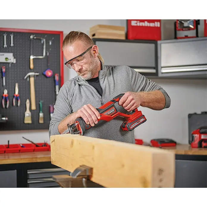Einhell PXC 18V Sierra de sable de batería TE-AP 18/26 Li - Solo - 18 V, Sin batería, 0 /min - 2.600 /min - Un hombre está usando una sierra de sable inalámbrica Einhell para cortar un trozo de madera, mientras usa gafas de seguridad.