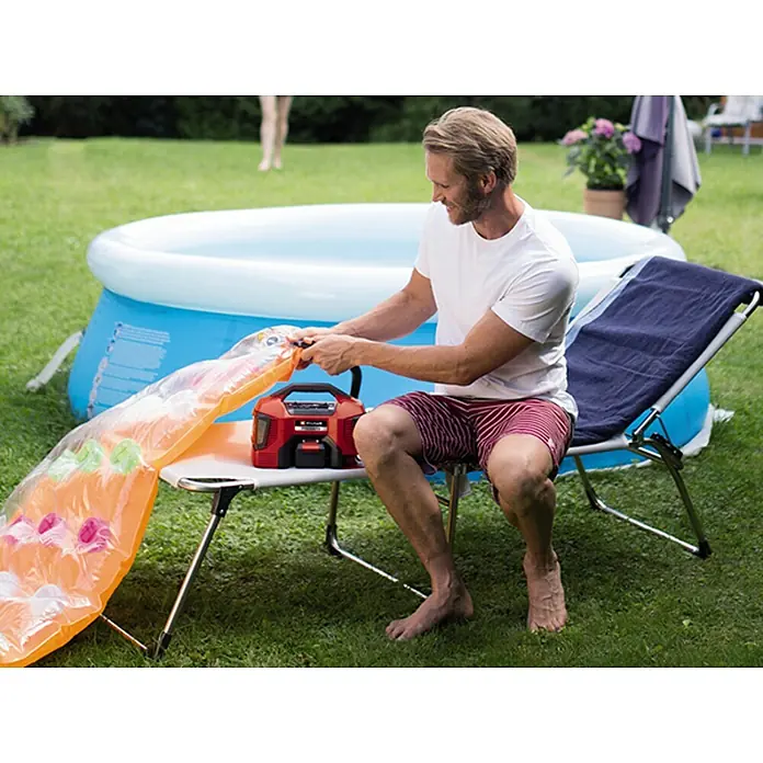 Einhell PXC 18V Compresor de batería PRESSITO 18/21 - 10,3 bar, 18 V, Sin batería - Un hombre sentado en una silla inflando un colchón de aire con una bomba Einhell roja, con una piscina al fondo.