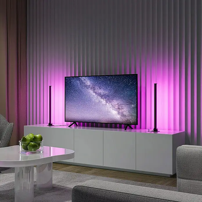 Trio Lighting Lámpara de sobremesa Boy - 2 ud., 5 W, RGBW - Dos barras de luz LED inteligentes junto a un televisor sobre un mueble blanco, creando una iluminación ambiental y colorida.