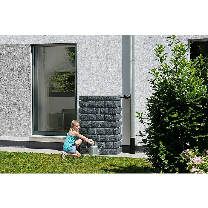 Garantia Exklusive Line Regenspeicher Rocky Junior - 300 l, Dark Granit - Kind füllt Gießkanne an einem grauen, blockartigen Wasserbehälter, der an einem Haus mit einem Fallrohr befestigt ist.