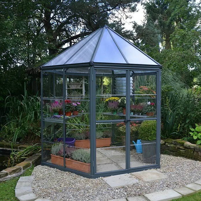 Palram – Canopia Gewächshaus Oasis - Außenmaß inkl. Dachüberstand (B x T): 213,2 x 247,1 cm, 0,7 mm, Anthrazit - Ein Gewächshaus mit Pflanzenregalen und einer klaren Dachkonstruktion. Es steht im Garten, umgeben von viel Grün und Blumen.