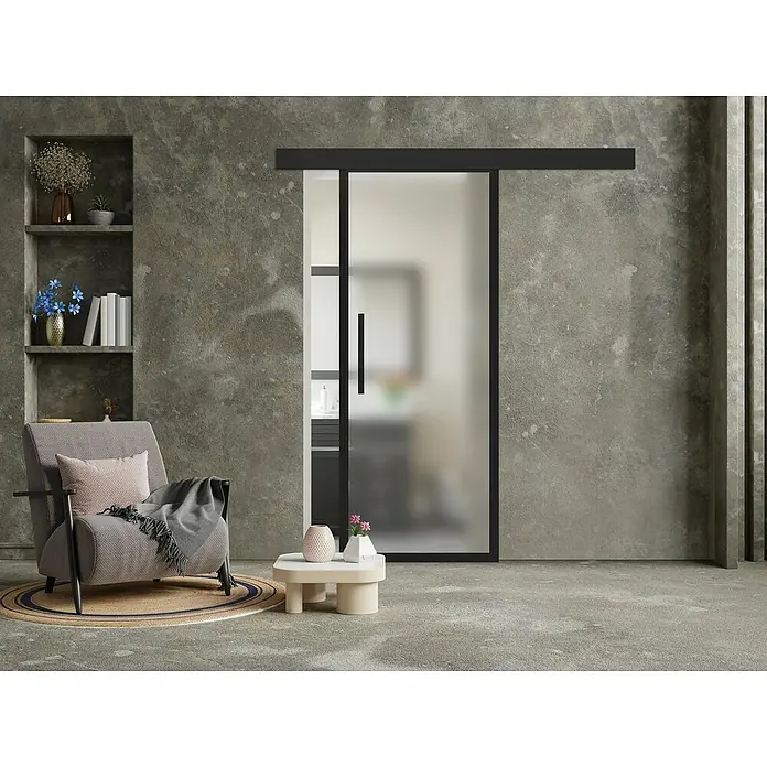 Puerta corredera de vidrio con herrajes Yumei - 95 x 210 cm, Vidrio templado, Herraje oculto - Un interior moderno con un rincón de lectura gris, una alfombra redonda y una puerta corredera de cristal con marco negro.