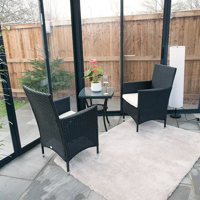 Palram – Canopia Pérgola de jardín cerrado Garda - L x An x Al: 5,95 x 5,17 x 3,13 m, Aluminio, Gris - Una acogedora habitación con dos sillas de mimbre negro y una pequeña mesa adornada con una planta de interior y una copa de vino.