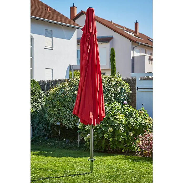 Sunfun Einschraub-Bodendübel - Ø x L: 6,5 x 51 cm, Stahl, Für Rohrdurchmesser: 38 mm - 58 mm - Ein roter, geschlossener Sonnenschirm steht auf einem Rasen, umgeben von üppigem Grün und Wohnhäusern. Praktischer Sonnenschutz für den Garten.