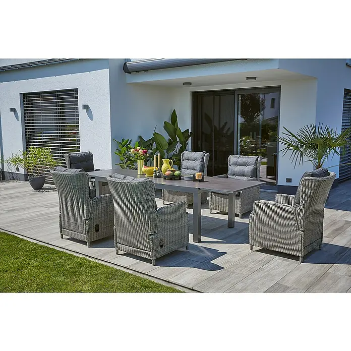 Sunfun Gartenmöbel-Set Maja Stone/Pauline - 7 -tlg., Sintered Stone/Aluminium/Polyrattan, Tischplatte ausziehbar, Anthrazit/Grau - Ein Gartentisch-Set mit sechs Rattansesseln und grauen Kissen steht auf einer Terrasse mit Pflanzen im Hintergrund.