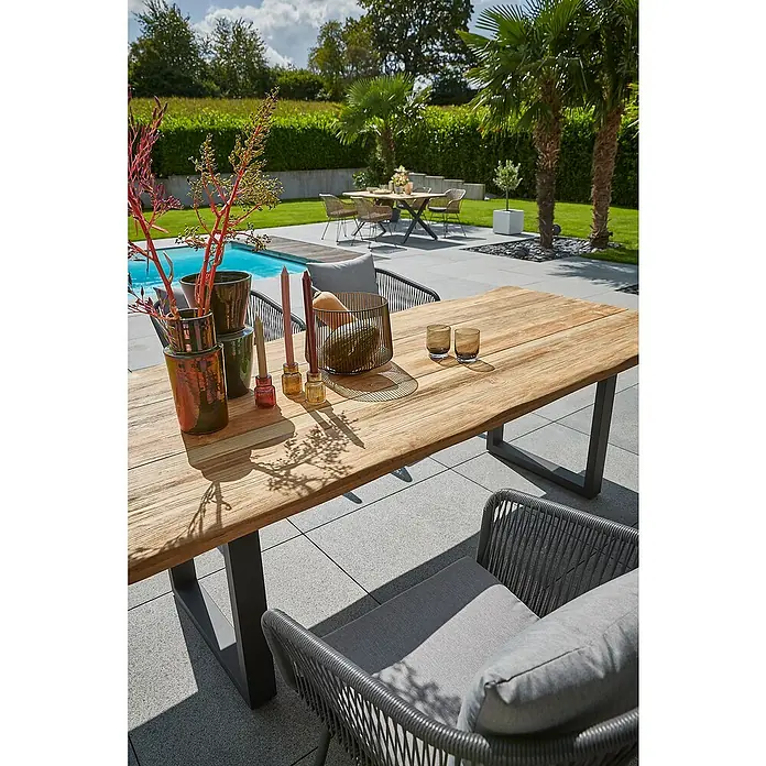 Sunfun Garten-Tischplatte180 x 90 cm, Teakholz, Baumkante - Ein rechteckiger Esstisch aus Teakholz im Freien mit Dekorationen, flankiert von grauen Korbstühlen, mit Pool und Garten im Hintergrund.