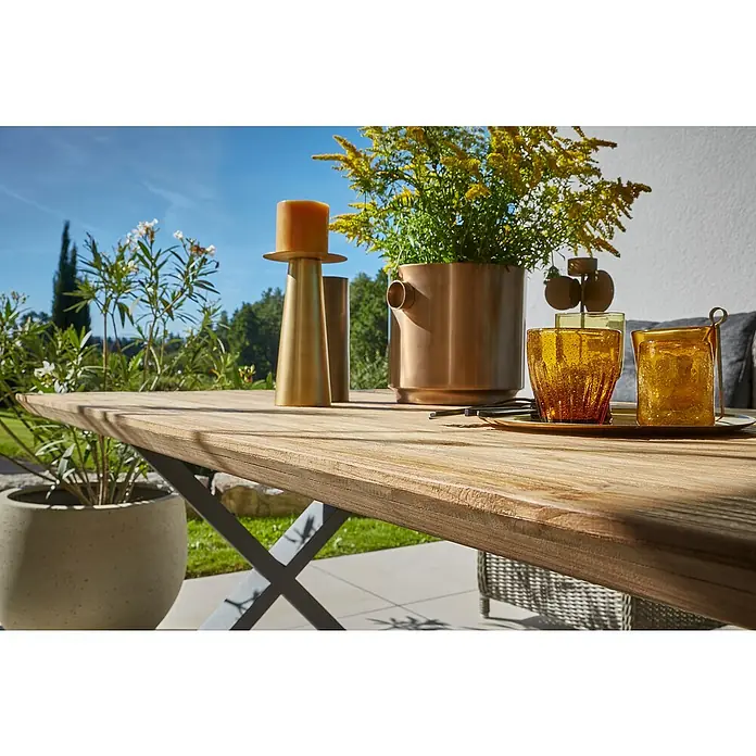 Sunfun Garten-Tischplatte - L x B: 180 x 90 cm, Teak, Schweizer Kante - Nahaufnahme eines Holztisches mit Kerze, Blumentopf und Gläsern im Freien. Einladung zum stilvollen Verweilen.