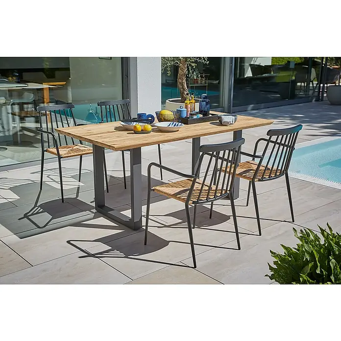 Sunfun Gartenmöbel-Set Eva - 6 -tlg., Teak/Edelstahl/Rattan, Teak/Grau/Naturbraun, Schweizer-Kante/U-Gestell - Holz Gartentisch mit sechs Stühlen. Der Tisch ist gedeckt und steht neben dem Schwimmbad im Garten.