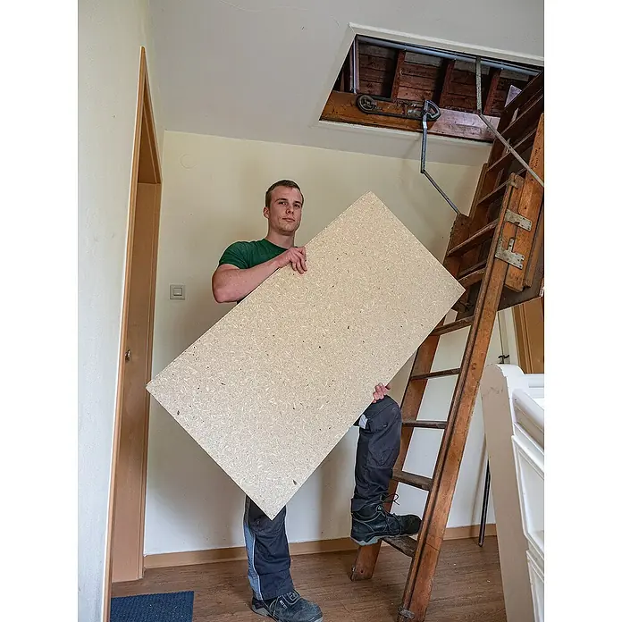 elka ESB-Platte RENO1.275 x 495 x 22 mm - Ein Mann trägt eine große Spanplatte vor einer Dachbodentreppe. Das Produkt ist die Spanplatte, ideal für Bau- oder DIY-Projekte.