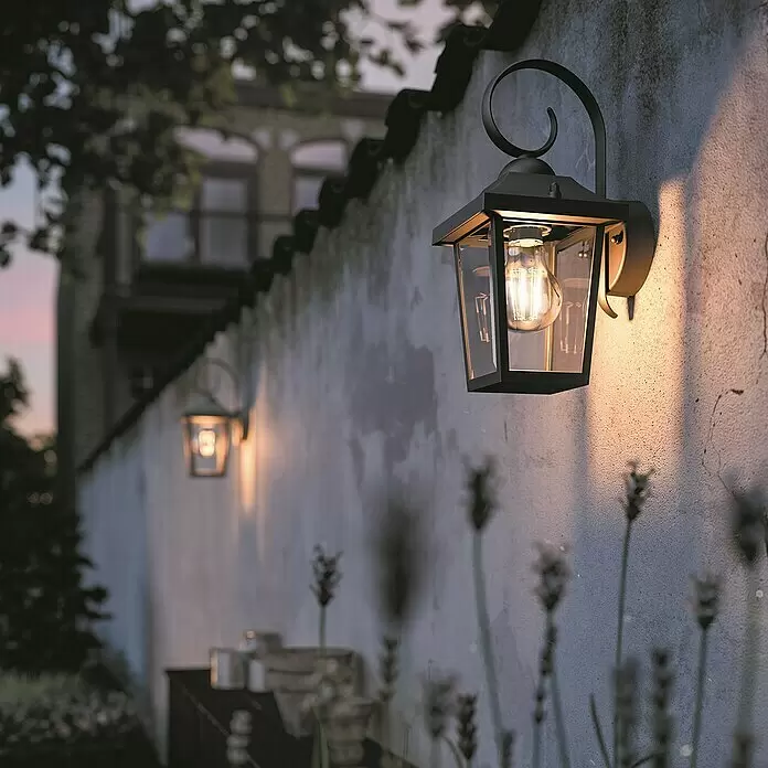 Philips Außenwandleuchte - 60 W, 14,3 x 17,8 x 30,8 cm, IP44 - Schwarze Outdoor-Wandleuchte aus Metall mit Klarglas und glühender Edison-Glühbirne an einer strukturierten Gartenmauer.