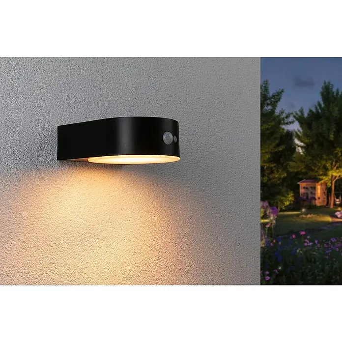 Paulmann Aplique solar para exterior Eileen - Potencia máx. del sistema: 5,5 W, L x An x Al: 19,8 x 17,5 x 6,5 cm, Negro, Flujo luminoso: 300 lm - Lámpara negra de exterior con sensor de movimiento montada en una pared gris, emitiendo una luz cálida.