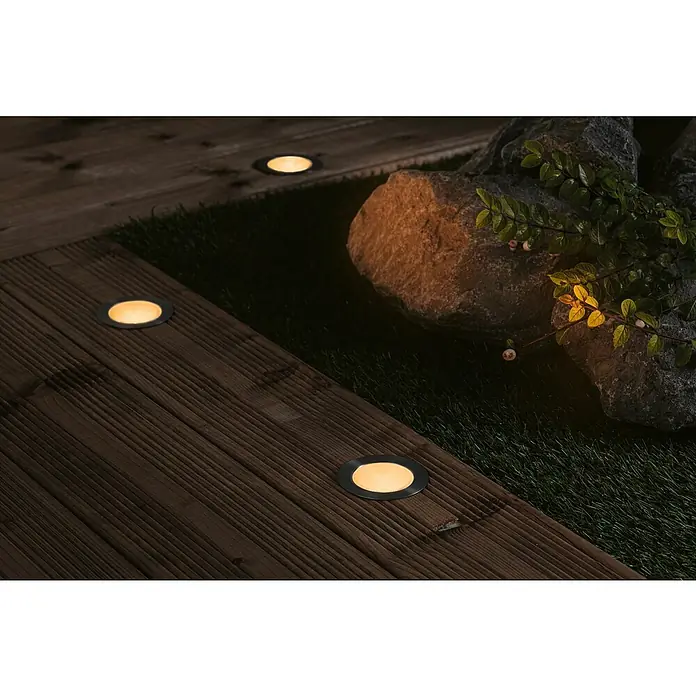 Paulmann Plug & Shine Foco para empotrar en suelo - 4,5 cm, Ámbar, IP67, Gris - Luces empotradas en el suelo iluminan un sendero de madera junto a piedras y césped, creando un ambiente suave para espacios exteriores.