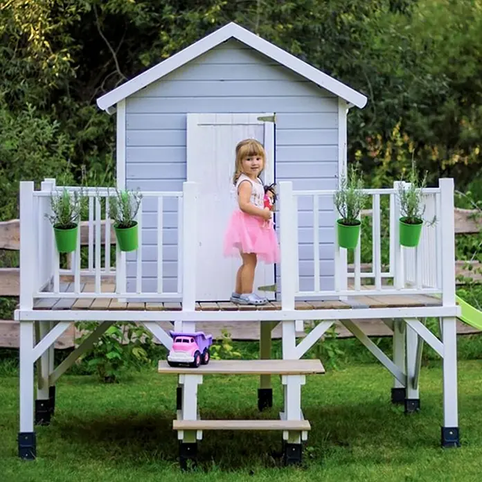 Spielhaus Mario - L x B x H: 227 x 286 x 232 cm, Holz, Natur - Ein Mädchen steht vor einem hölzernen Spielhaus, das auf einer Plattform mit Geländer steht, mit dekorativen Pflanzgefäßen und einem kleinen Tisch darunter.