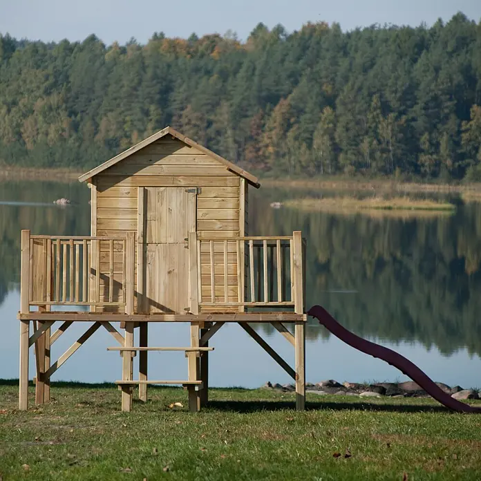 Spielhaus Mario - L x B x H: 227 x 286 x 232 cm, Holz, Natur - Ein hölzernes Spielhaus mit Rutsche steht auf einer grünen Wiese vor einem See und Bäumen im Hintergrund.