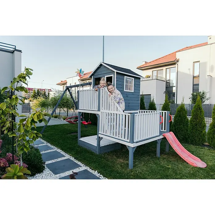 Spielhaus Tom 60 - L x B x H: 226,5 x 176 x 272 cm, Holz, Natur/Rot - Ein zweistöckiges Holzspielhaus mit Rutsche, Schaukel und Veranda, umgeben von Rasen und Pflanzen, bietet Kindern einen sicheren und unterhaltsamen Spielplatz.