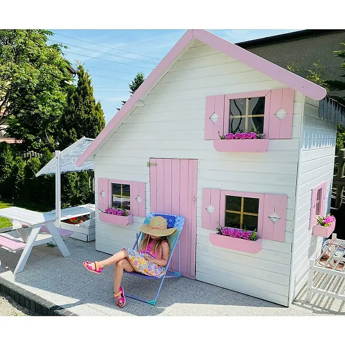 Spielhaus Aleks - L x B x H: 223 x 250 x 230 cm, Holz, Natur - Mädchen entspannt vor einem weißen Spielhaus mit rosa Akzenten, das mit Blumenkästen dekoriert ist.