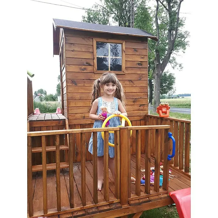 Spielhaus Tom - L x B x H: 226,5 x 176 x 272 cm, Holz, Natur - Ein lächelndes Mädchen steht auf einem hölzernen Spielplatz mit Lenkrad und Puppe, vor einem kleinen Holzhaus.