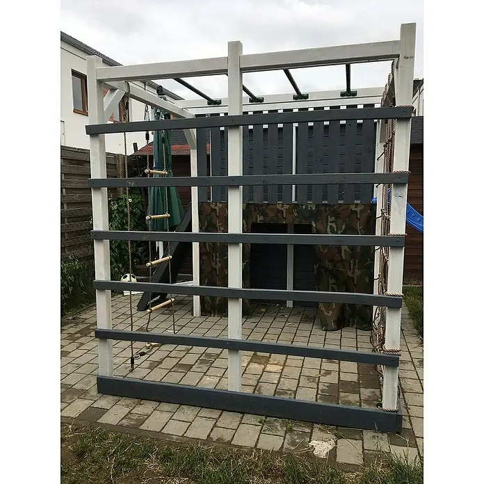 Kinderspielturm Kubisch Alfa - L x B x H: 447 x 225 x 210 cm, Natur - Ein hölzerner Spielplatz mit Klettergerüst, Strickleiter und Tarnvorhang steht auf einem gepflasterten Bereich im Garten. Ideal für Kinder zum Austoben!