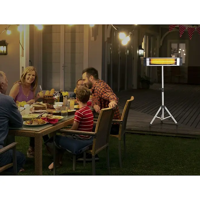 Purline Estufa de pie infrarrojos Heaty EXC 30 - 10 x 101 x 180 cm, 3.000 W - Un calentador de patio sobre un trípode está al lado de una familia cenando al aire libre, proporcionando calor para una velada acogedora.