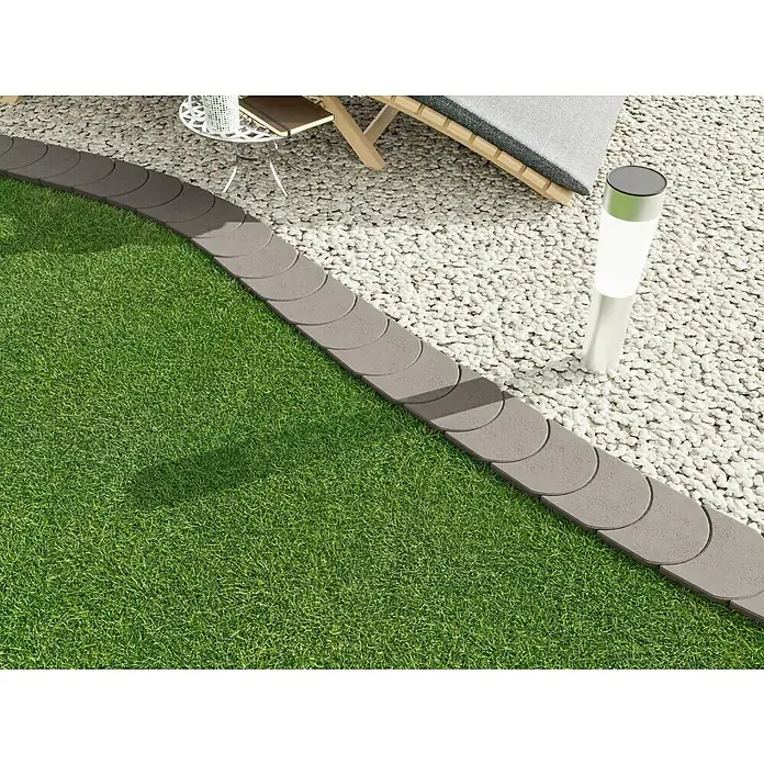 Christoph Mähstein Mähroboter-Randplatte - 15 x 24 x 4,2 cm, Grau, Beton - Rasenkante aus Beton mit gerundeten Kanten, die einen Rasen von einem Kiesweg trennt, und eine Solarleuchte im Hintergrund.