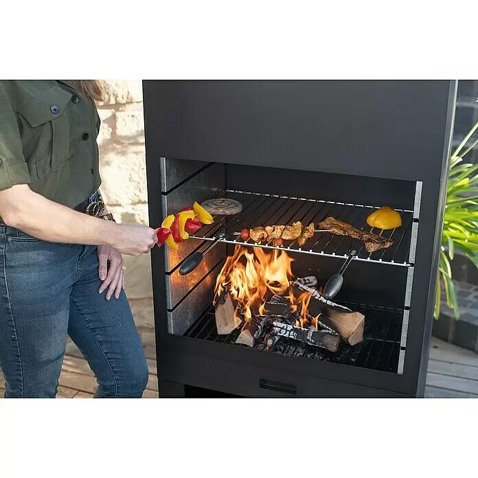Westmann Grillkamin - L x B x H: 410 x 700 x 2.290 mm, Schwarz, Stahl - Schwarzer Outdoor-Grill mit Holzfeuer, auf dem Fleisch, Wurst und Gemüsespieße zubereitet werden. Eine Person hält einen Spieß.