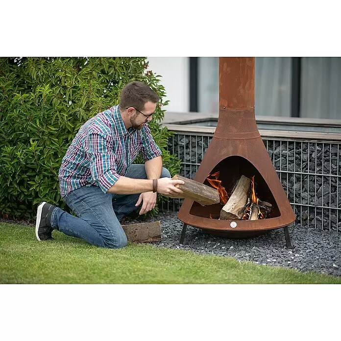 Westmann Feuerstelle - 70 x 70 x 160 cm, Metall, Rost - Rostfarbene Metall-Feuerstelle mit einem Mann, der Holz ins brennende Feuer legt, im Garten.
