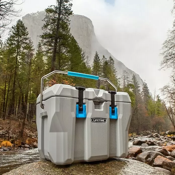 Lifetime Kühlbox Cooler - 26,5 l, Grau/Blau - Graue Lifetime Kühlbox mit blauem Griff und Verschlüssen steht auf einem nassen Felsen am Fluss, mit nebligen Bergen und Bäumen im Hintergrund.