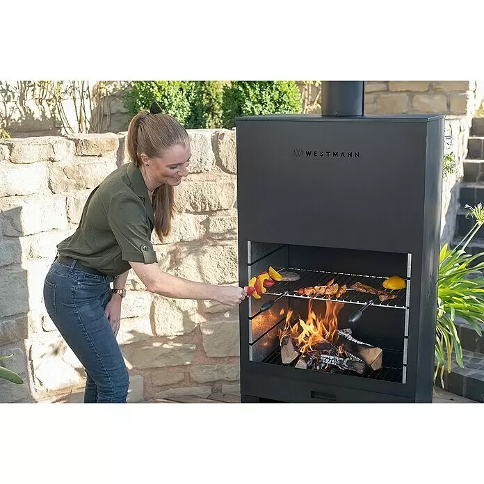 Westmann Grillkamin - L x B x H: 410 x 700 x 2.290 mm, Schwarz, Stahl - Schwarzer WESTMANN Outdoor-Grill, eine Frau legt Spieße auf den Rost, Feuer brennt darunter.