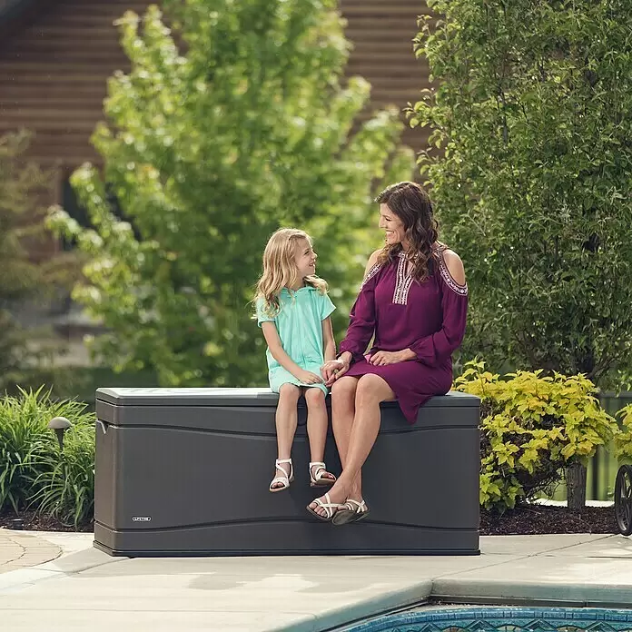 Lifetime Auflagenbox - 573 x 1.525 x 67 cm, Kunststoff, Anthrazit - Dunkelgraue Outdoor-Aufbewahrungsbox, die als Bank am Pool genutzt wird, mit einer Frau und einem Kind darauf sitzend.