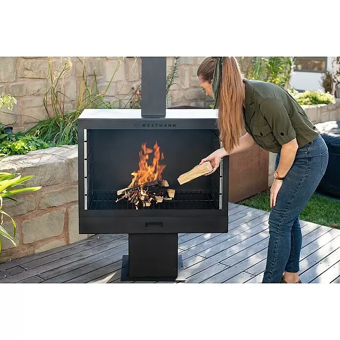 Westmann Grillkamin - L x B x H: 500 x 300 x 1.600 mm, Stahl, Schwarz - Frau legt Holz in schwarzen Außenkamin/Grill mit Feuer auf Holzterrasse.