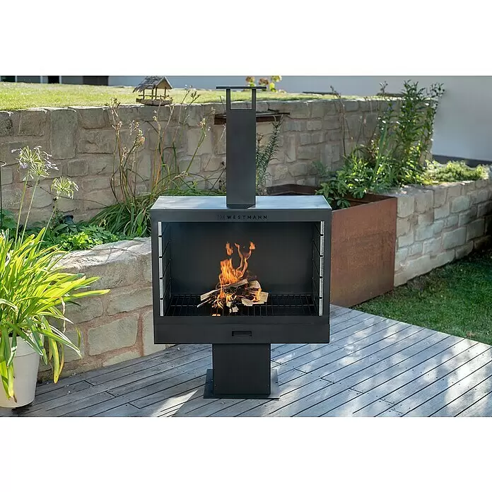 Westmann Grillkamin - L x B x H: 500 x 300 x 1.600 mm, Stahl, Schwarz - Schwarze Metall-Feuerstelle und Grill mit brennendem Feuer, auf einer Holzterrasse im Garten.