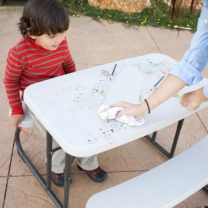 Lifetime Kinder-Picknicktisch - L x B x H: 83 x 90 x 54 cm, Polypropylen, Lichtgrau/Schwarz - Ein Junge steht an einem weißen Kindertisch mit Farbflecken, während eine Hand ihn mit einem Tuch abwischt.