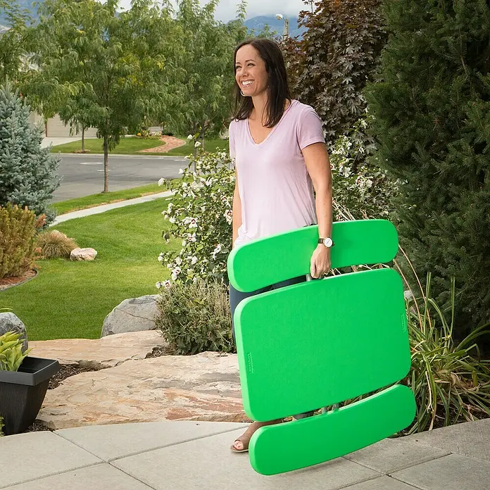 Lifetime Kinder-Picknicktisch - L x B x H: 83 x 90 x 54 cm, Polypropylen, Grün - Eine Frau trägt einen zusammengeklappten, grünen Kindertisch der Marke Lifetime im Freien vor einem Haus.