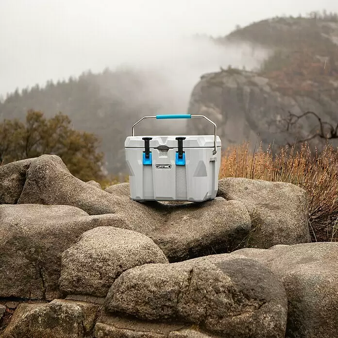 Lifetime Kühlbox Cooler - 26,5 l, Grau/Blau - Graue Kühlbox mit blauem Griff und Riegeln, beschriftet mit „LIFETIME 28“, auf Felsen in nebliger Naturlandschaft.