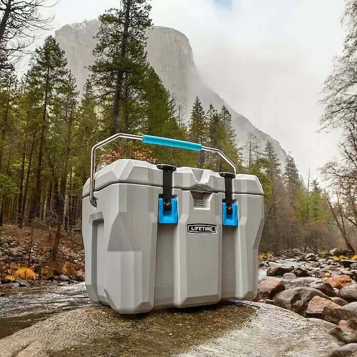 Lifetime Kühlbox Cooler - 26,5 l, Grau/Blau - Eine graue Kühlbox mit blauem Griff und Verschlüssen steht auf einem Felsen am Fluss vor Bäumen und einem nebligen Berg, was den Außeneinsatz andeutet.