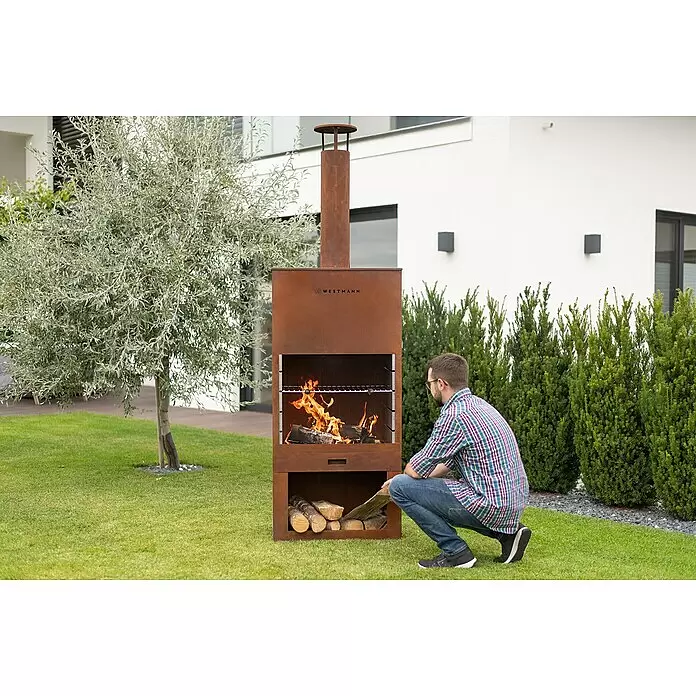 Westmann Grillkamin - L x B x H: 410 x 700 x 2.290 mm, Rost, Stahl - Corten Stahl Außenkamin/Grill, Mann legt Holz nach. Feuer brennt, zeigt funktionellen Einsatz im Garten.