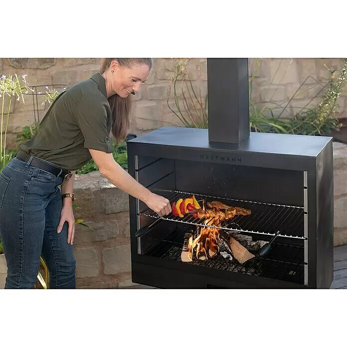 Westmann Grillkamin - L x B x H: 500 x 300 x 1.600 mm, Stahl, Schwarz - Schwarzer Westmann Outdoor-Grill, eine Frau legt Spieße auf den Rost über Holzfeuer.