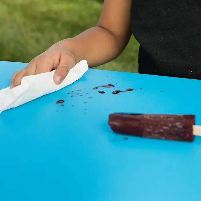 Lifetime Kinder-Picknicktisch - L x B x H: 83 x 90 x 54 cm, Polypropylen, Blau - Nahaufnahme eines Kindes, das einen blauen Tisch mit einem Papiertuch und restlichem Eis reinigt.
