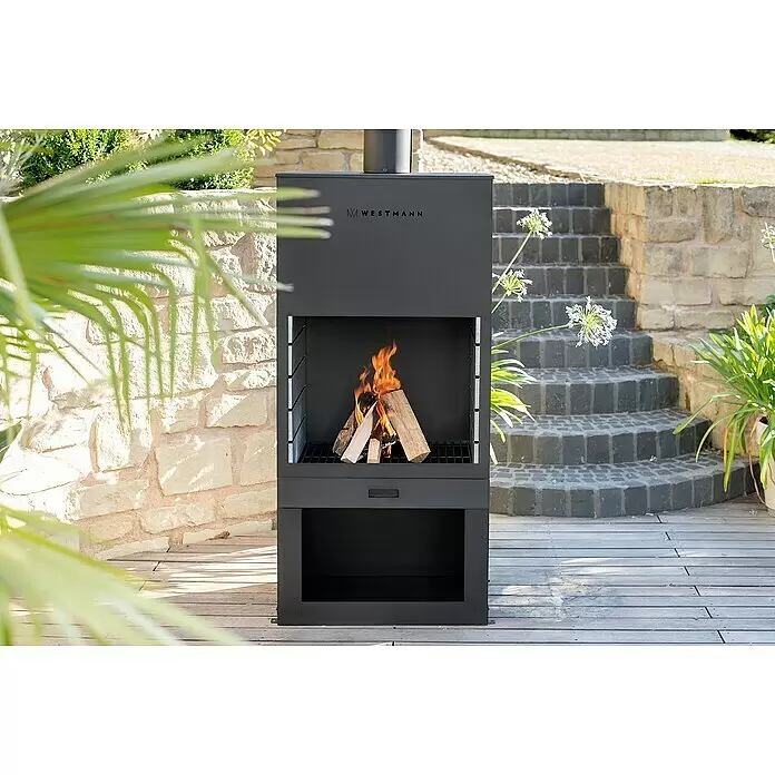 Westmann Grillkamin - L x B x H: 410 x 700 x 2.290 mm, Schwarz, Stahl - Dunkelgrauer Metall-Gartenkamin mit brennendem Feuer auf einer Holzterrasse. Mit Grillrost und Stauraum.